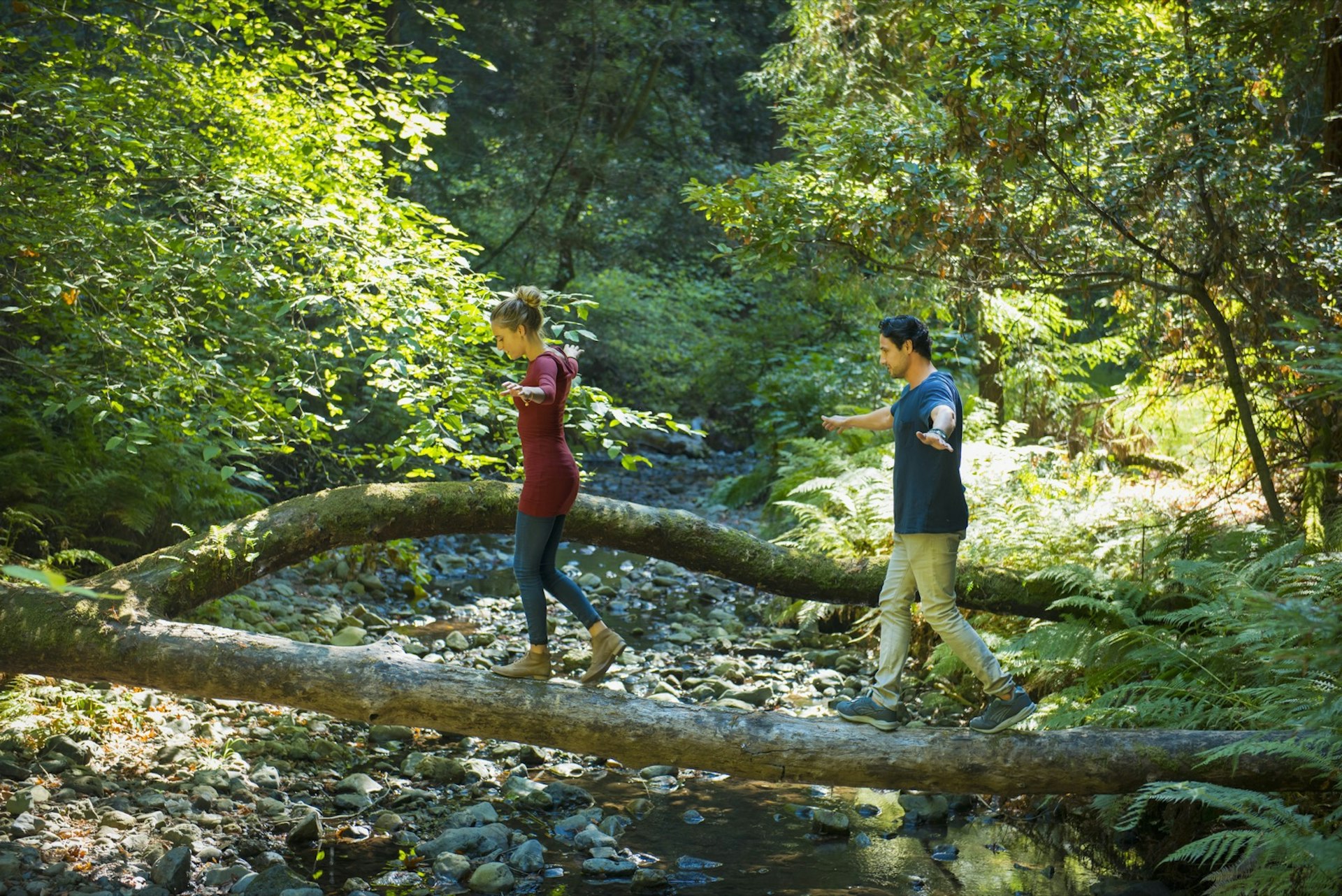 A man and a woman balance on a log across a shallow creek surrounded by green bushes