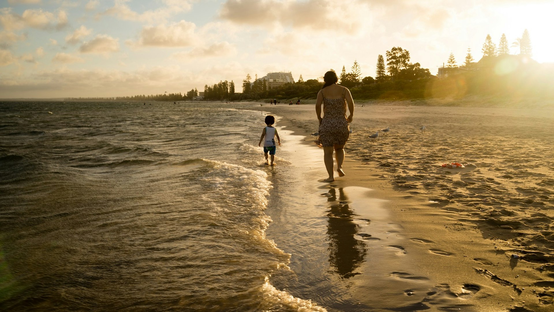 A parent and toddler are silhouetted as they walk on the edge of the sea away from the camera