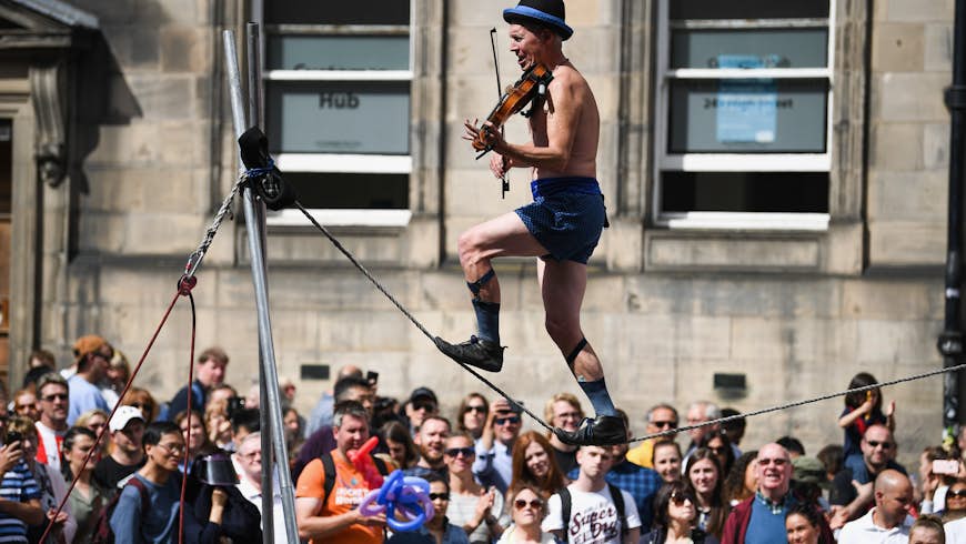 Edinburgh Festival Fringe entertainers perform on the Royal Mile. The Fringe is the largest performing arts festival in the world, with an excess of 30,000 performances of more than 2000 shows. A man is walking along a high line wearing shorts and a bowler hat while playing a violin. A huge crowd is gathered around to watch. 