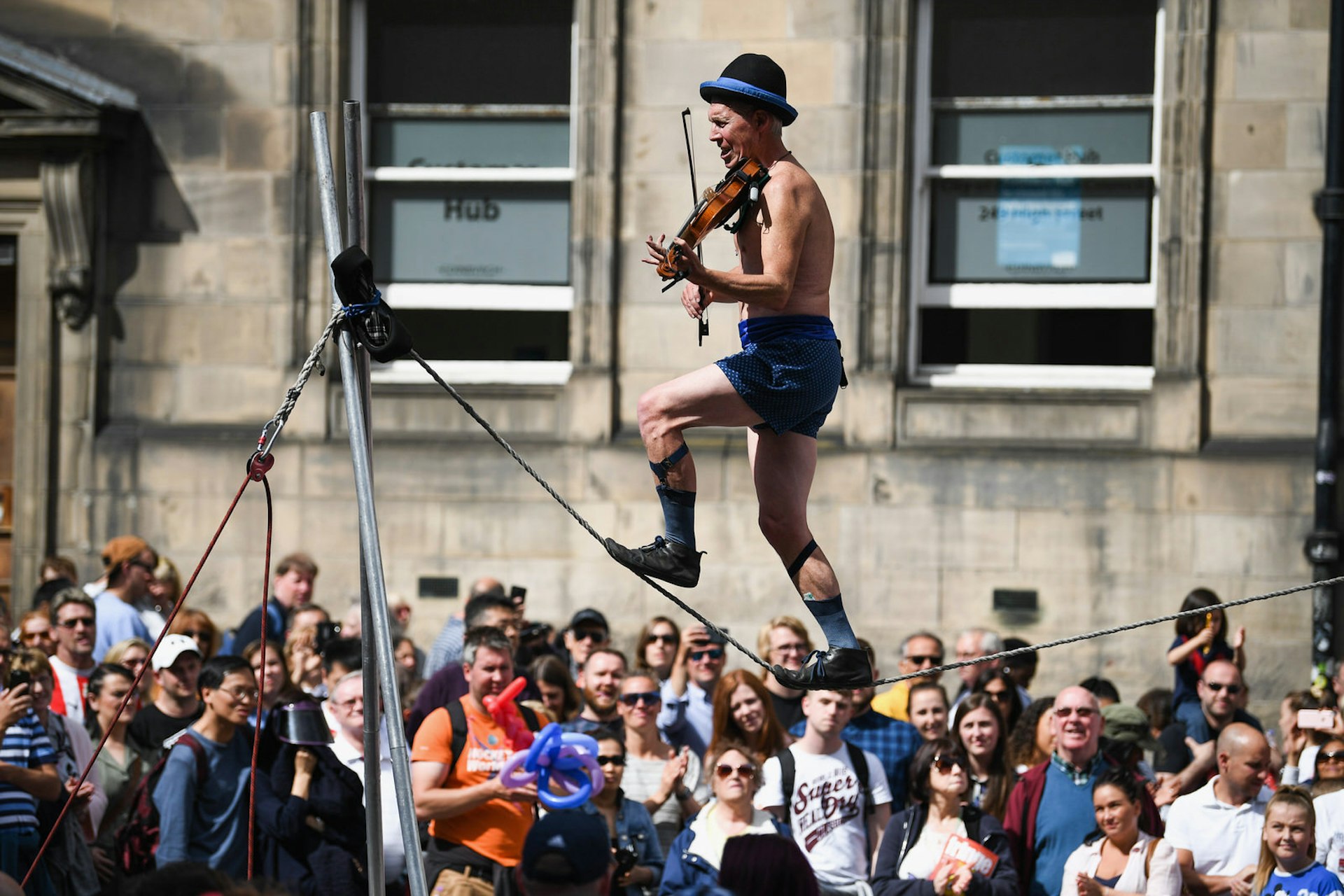 Edinburgh Festival Fringe entertainers perform on the Royal Mile. The Fringe is the largest performing arts festival in the world, with an excess of 30,000 performances of more than 2000 shows. A man is walking along a high line wearing shorts and a bowler hat while playing a violin. A huge crowd is gathered around to watch. 