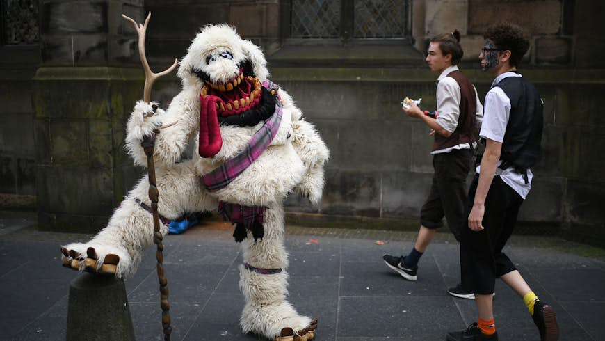 Edinburgh Festival Fringe entertainers perform on the Royal Mile. A person in an oversized white yeti costume with a tartan sash and holding a staff props their leg up on a bollard while two men walk by. 