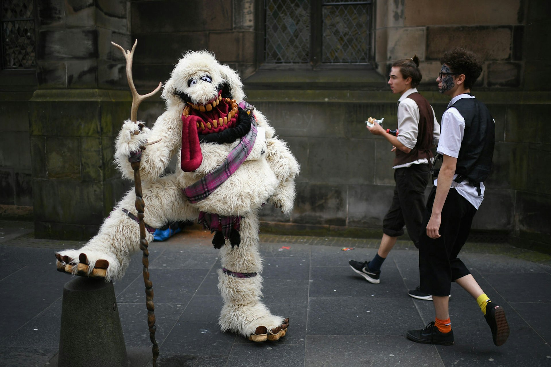 Edinburgh Festival Fringe entertainers perform on the Royal Mile. A person in an oversized white yeti costume with a tartan sash and holding a staff props their leg up on a bollard while two men walk by. 