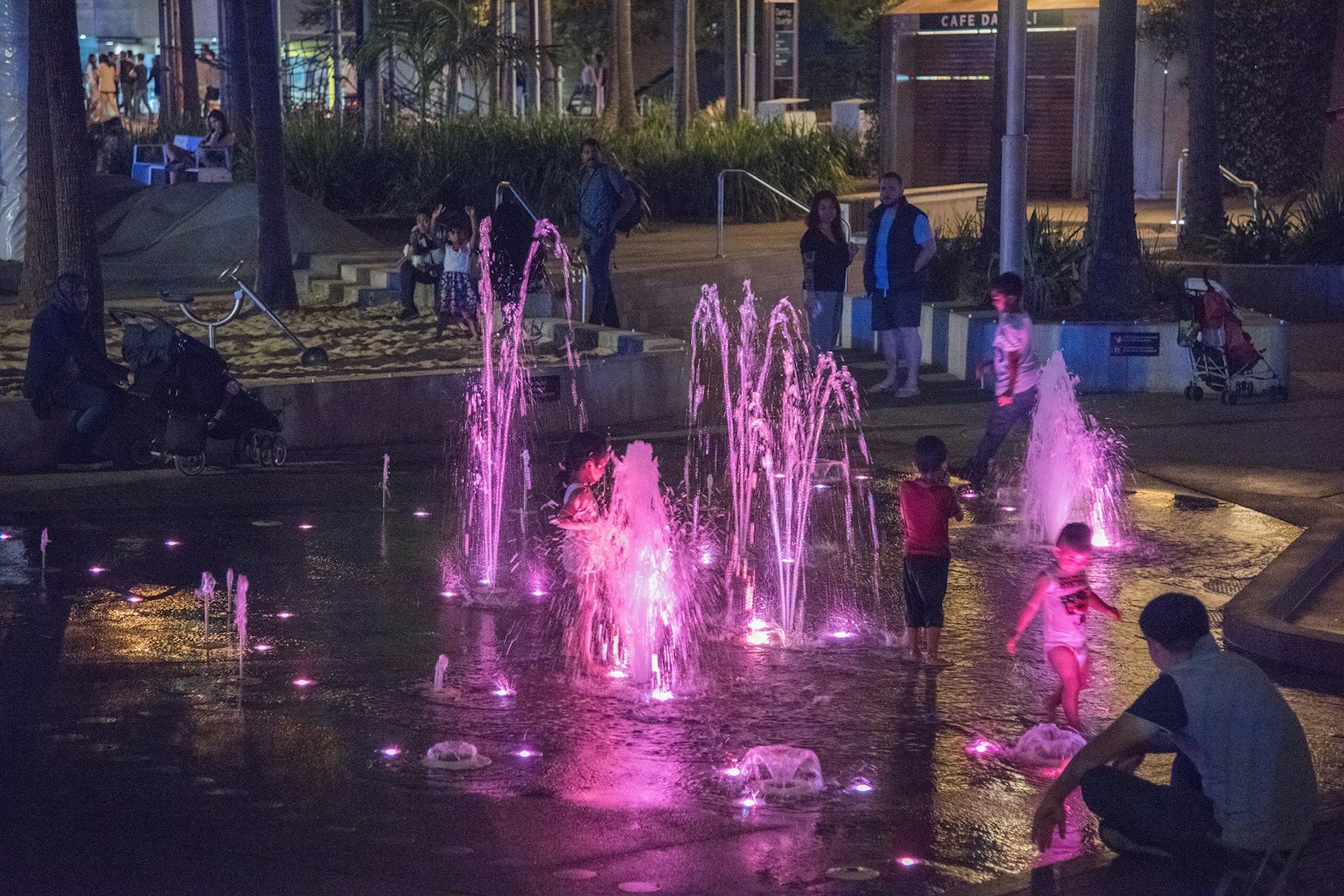Several jets of water are illuminated pink in the darkness as three children play in the fountain