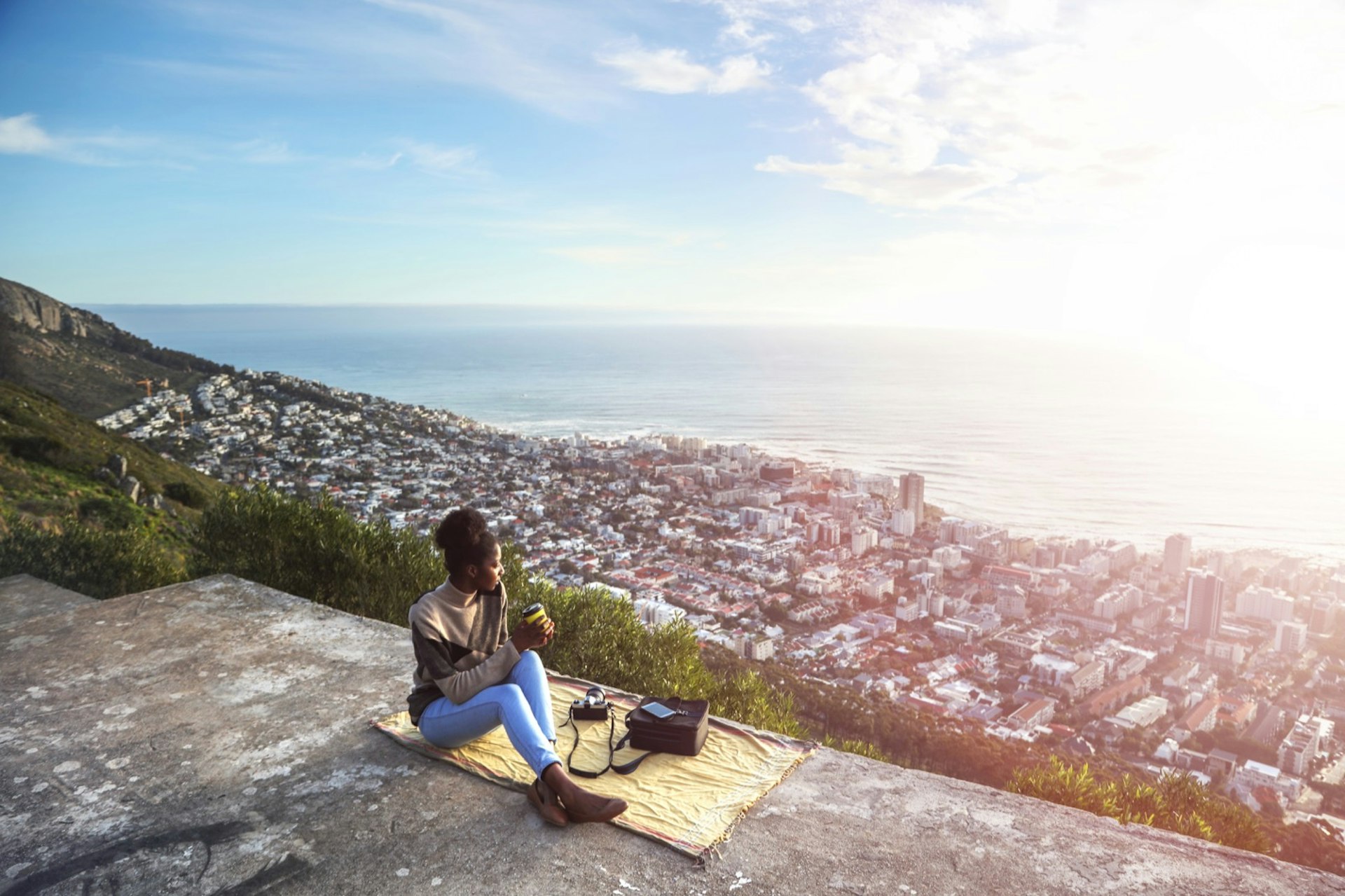 A woman sits on a yellow towel on a hill overlooking a large city on the coast; sacred spaces 