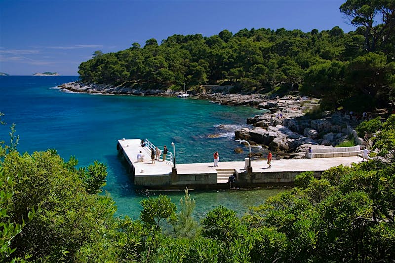 A weekend in Dubrovnik should involve a trip to Lokrum; we see the harbour, with a few tourists waiting for the next ferry; beyond is a stony seashore, with thickly wooded trees behind it.