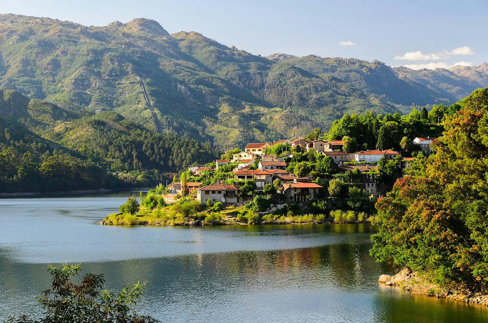 A view of a village clinging to a hill rising from the banks of the Cavado river in Peneda-Gerês national park in northern Portugal, with rugged green hills beyond.