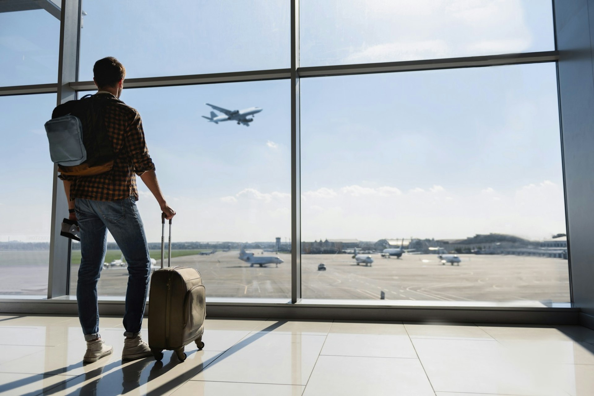 Young man is standing near window at the airport and watching plane before departure. He is standing and carrying luggage. Focus on his back; flying etiquette