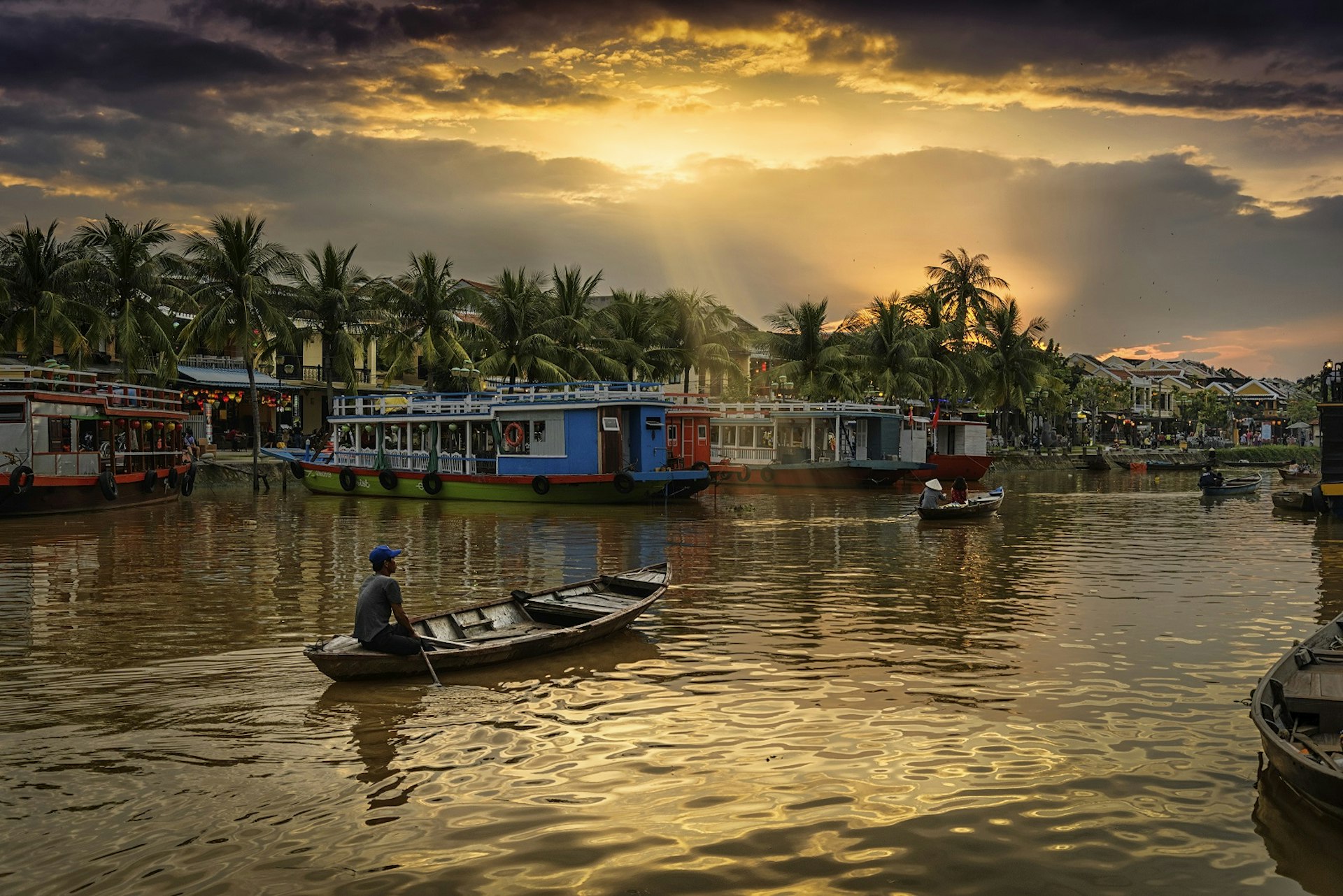 A few small wooden boats on the Thu Bon river with the sun setting over palm trees. There are big, colourful boats docked along the banks and restaurants beyond them. 
