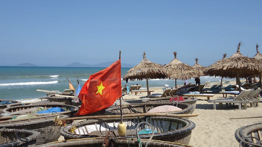 A Vietnamese flag flies above a basket boat on the beach, with some sunshades and sunloungers in the background
