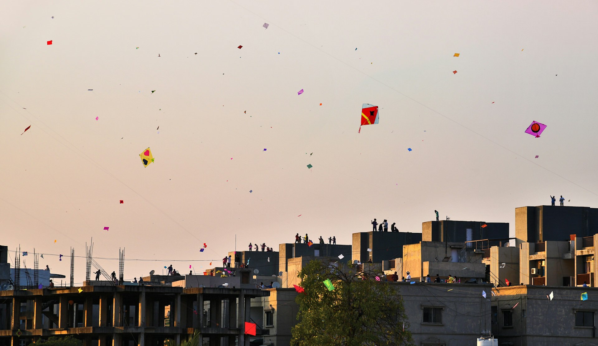 Colourful kites fly above the rooftops of Ahmedabad at dusk. Silhouettes of people are visible on the top of buildings.