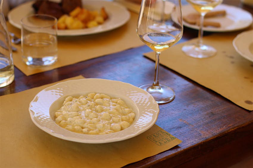 A plate of creamy gnocchi served on a white plate with other dishes, glasses of water and white wine blurred in the background on a dark wooden table