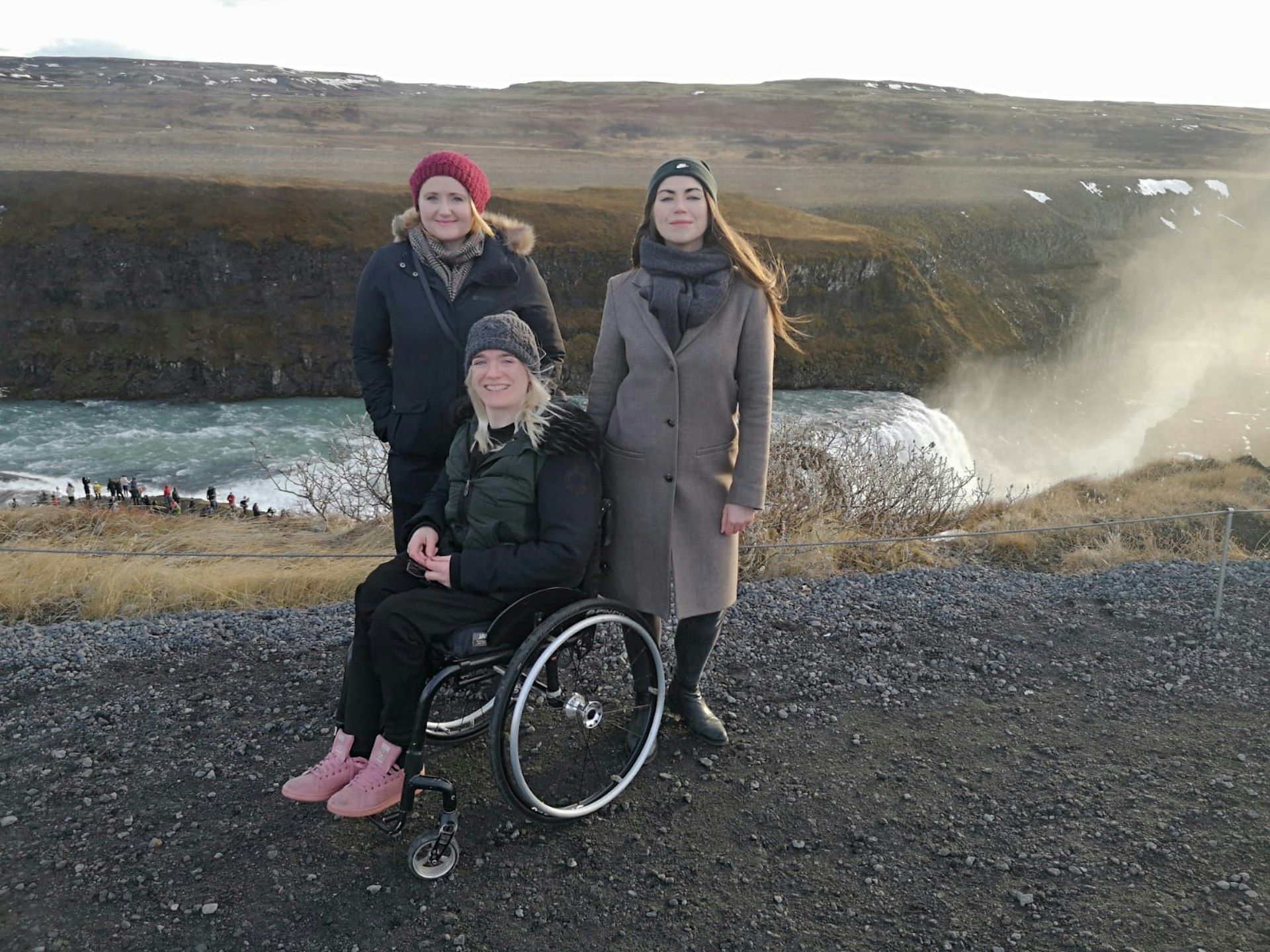Three women are standing on a gravel path next to the top of a waterfall. The woman in the centre is in a wheelchair and her two friends are standing behind her.
