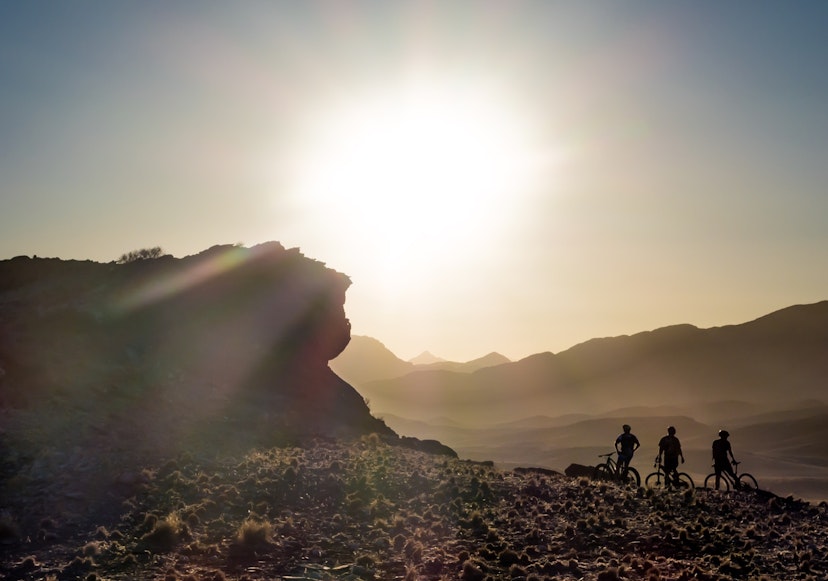 The silouette of three people on bicycles next to a large rock in front of a setting sun