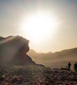 The silouette of three people on bicycles next to a large rock in front of a setting sun