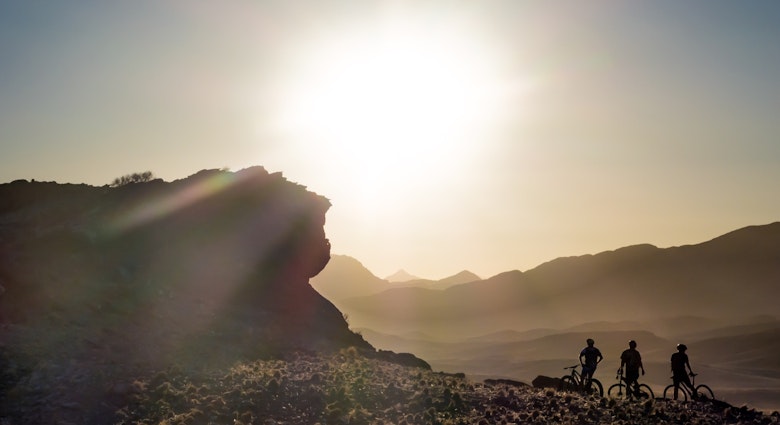 The silouette of three people on bicycles next to a large rock in front of a setting sun