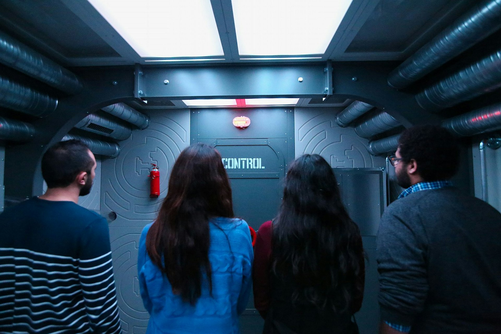 Four people stand with their backs to the camera facing a door that says "Control" in a grey industrial-looking room; the ceiling is covered with strip lights and silver pipes.