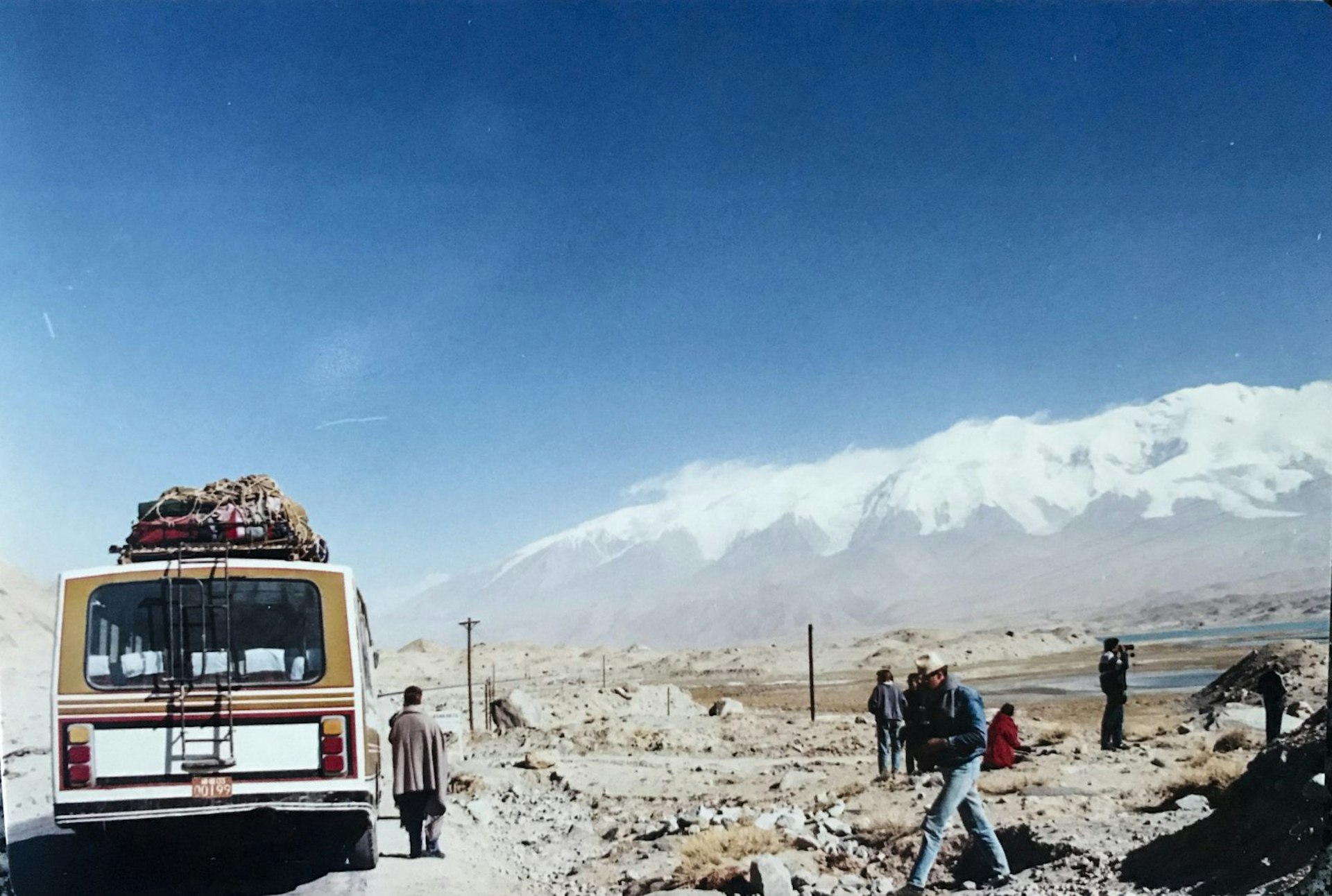 A bus is stopped on a remote road on the left of the picture. People are standing around taking pictures on the rocky ground to the right. Snow capped mountains loom in the back of the photo and the sky is blue and cloudless.