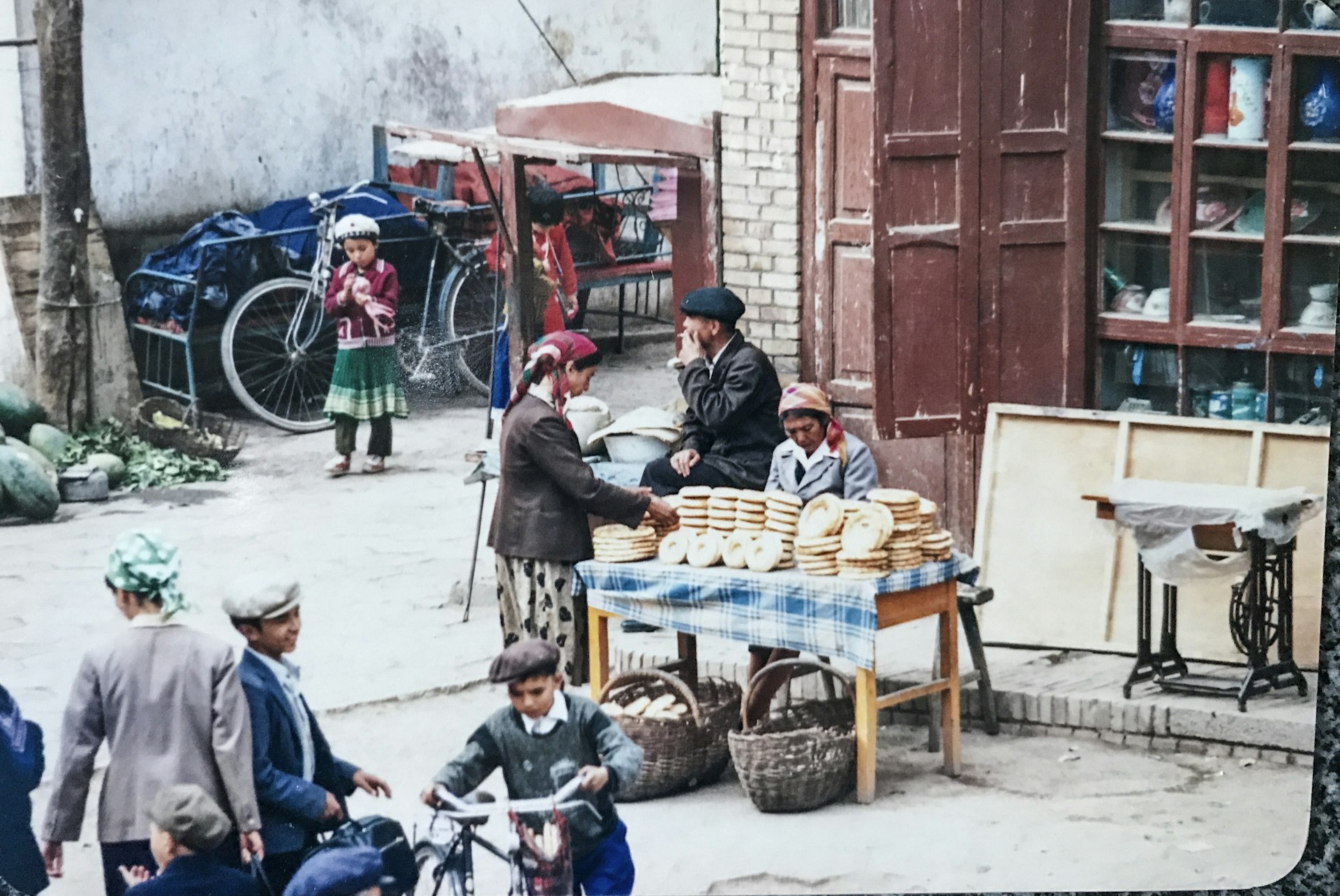 A table is set up on the side of the street and is covered in flat bread. A woman is looking at the bread on display while a child plays in the background and two young boys are on their bikes in the foreground. The stall is set up in front of a building with several windows.