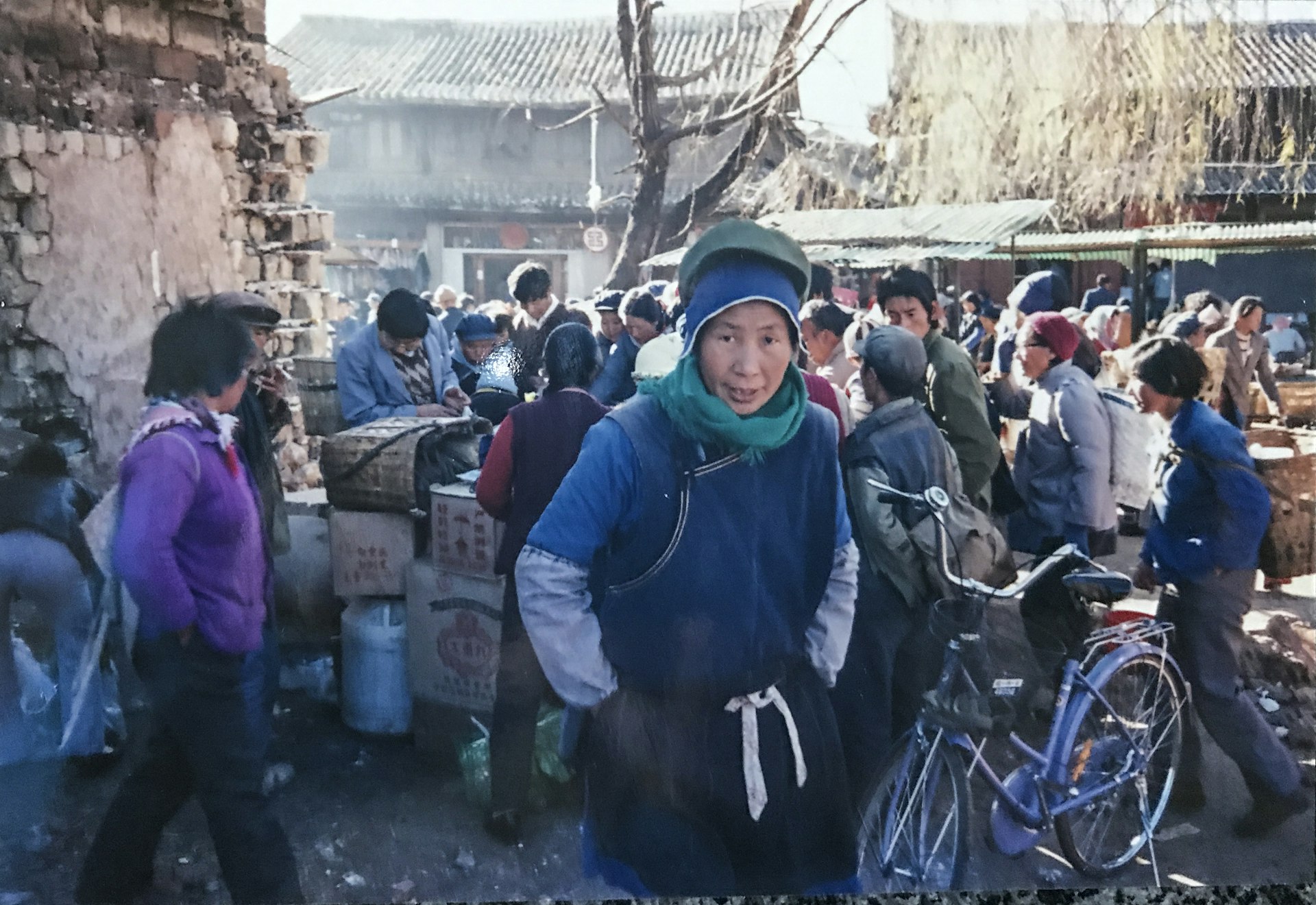 A scene of the heavily crowded market area. A Chinese woman in a blue jumper and hat, and black apron, stands in the foreground staring at the camera.