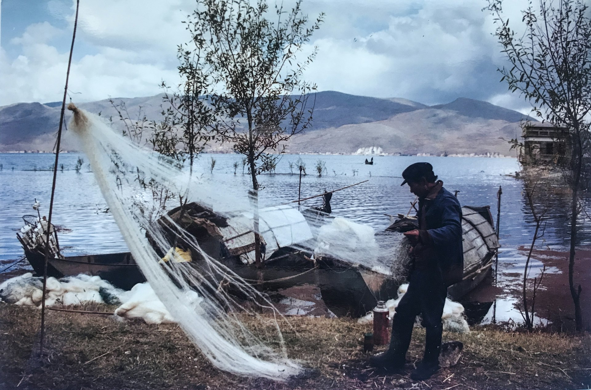 A fisherman detangles his nets on the banks of a river. He's wearing an overcoat and a black cap. The sky is blue and scattered with white clouds and the other bank of the river is in the background of the image. Hilly peaks frame the river. Two boats are moored on the bank next to the fisherman.