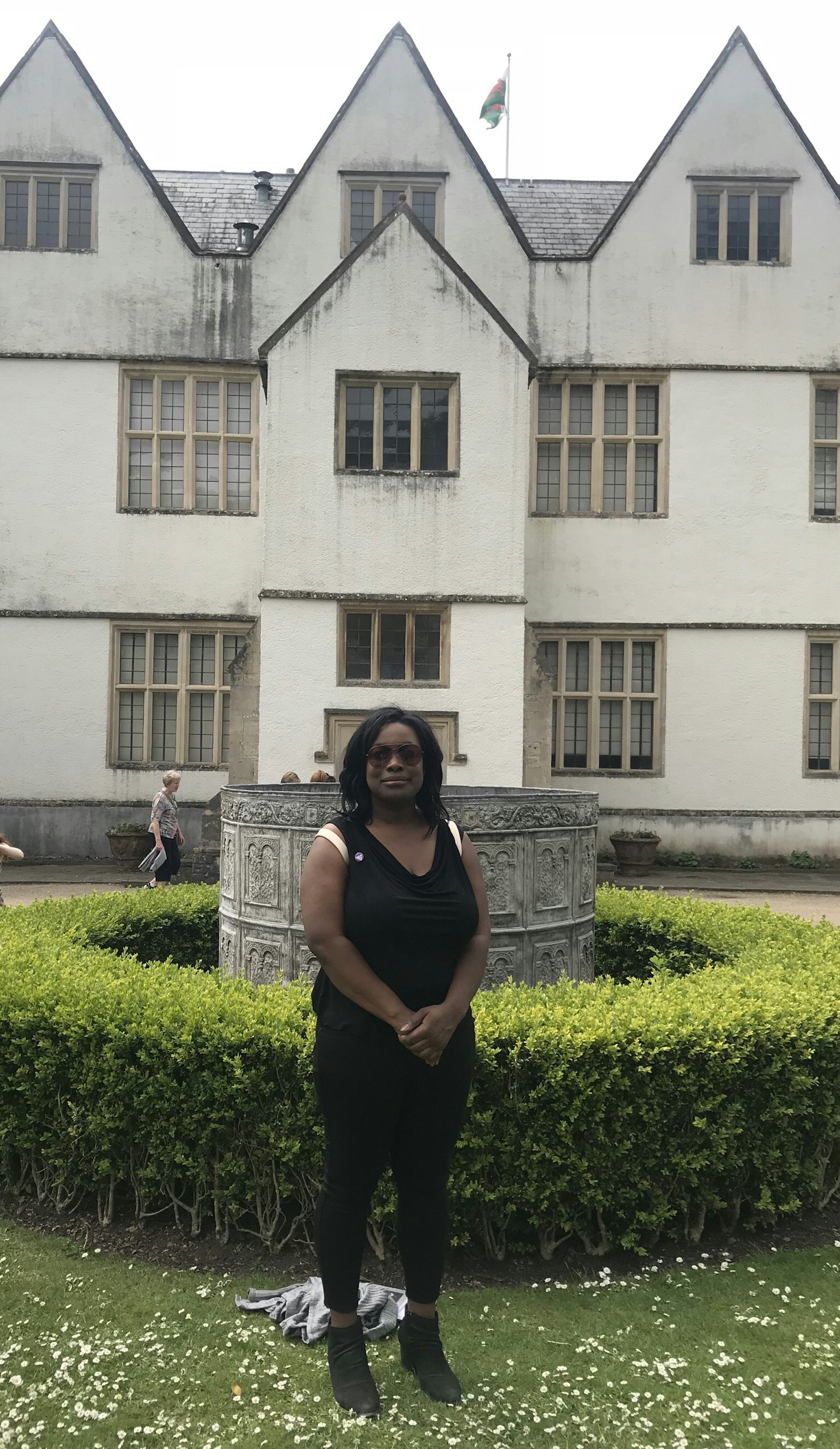 An African American woman stands in the garden at the from of St. Fagan's National Museum of History in Wales. She is wearing all black and sunglasses, standing in front of a decorative stone structure, surrounded by a green hedge. The museum in the background is painted a pale yellow, has a peaked roof and a welsh flag flying.