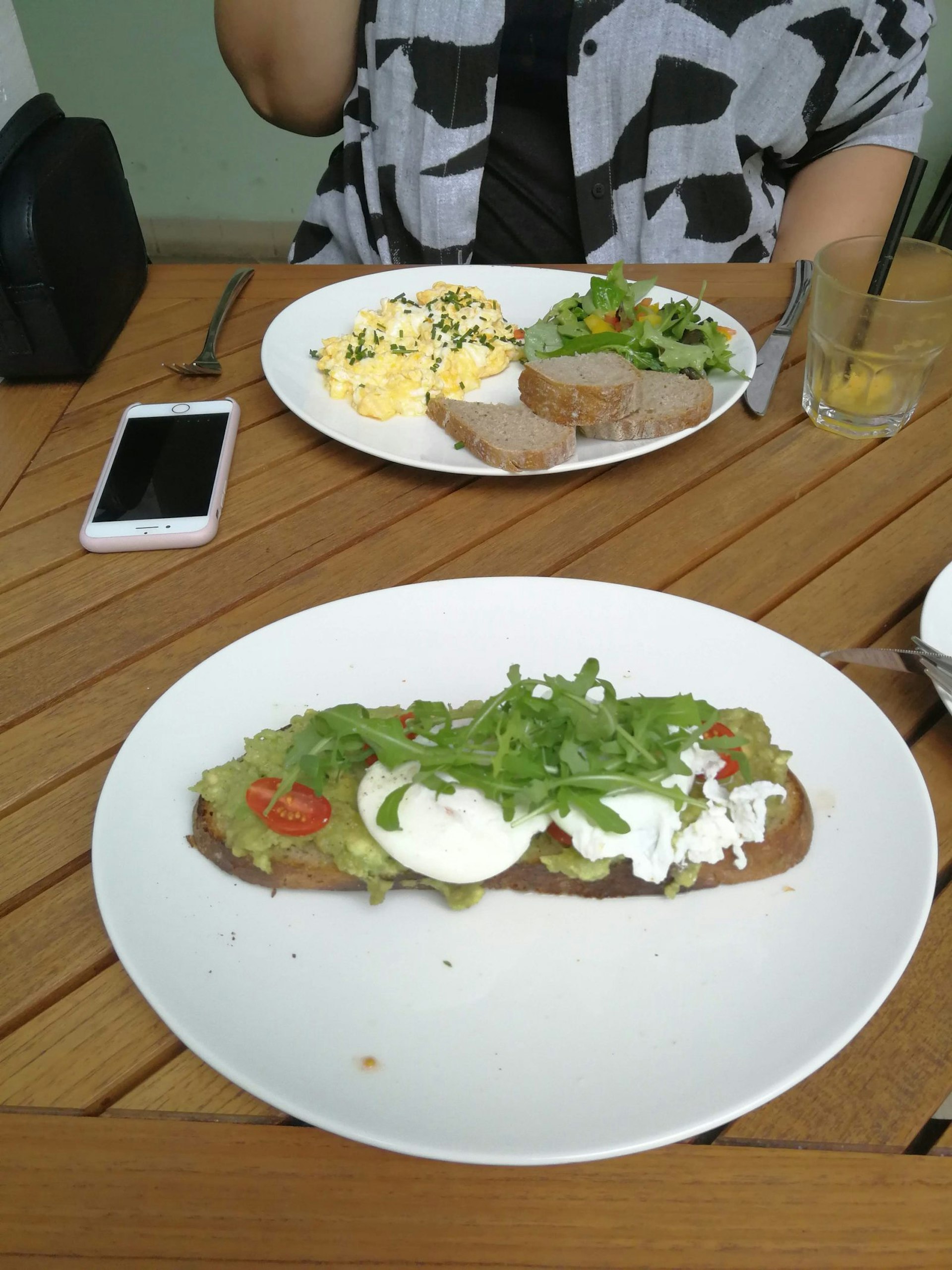 Two plates on a table: the one in the foreground has avocado and poached eggs on toast topped with arugula. The one in the background has scrambled eggs, bread and salad.