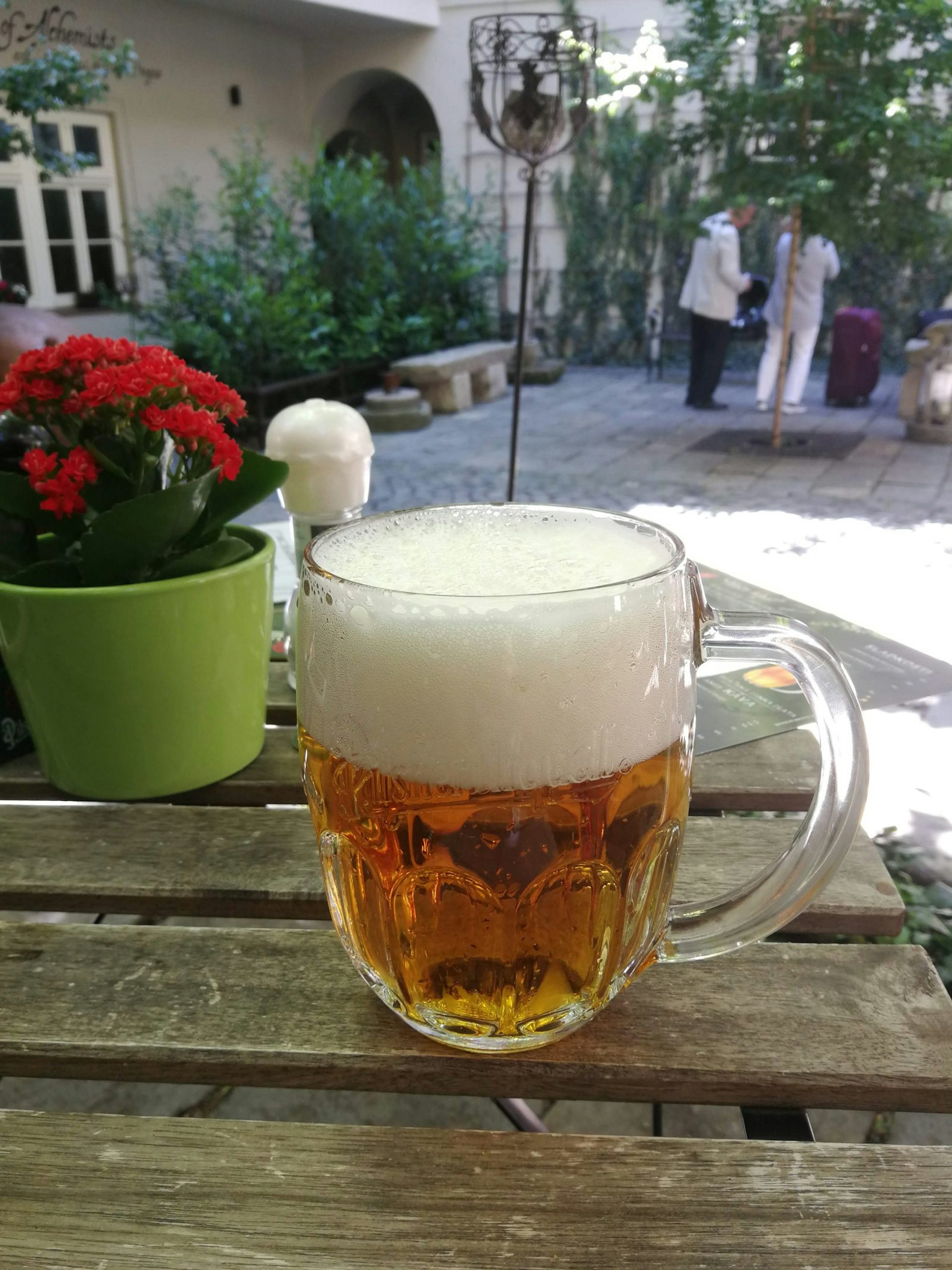 A pint of beer atop a picnic table in a pub garden. A flower pot sits behind the glass and two people stand across the yard.