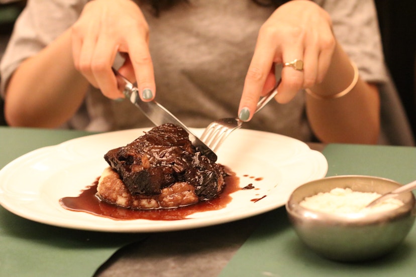 A cut of slow-cooked beef with a red wine sauce served on mashed potato. The meal is on a white plate and the close-up shot shows someone's hands cutting into the meat with silver cutlery.