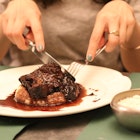 A cut of slow-cooked beef with a red wine sauce served on mashed potato. The meal is on a white plate and the close-up shot shows someone's hands cutting into the meat with silver cutlery.