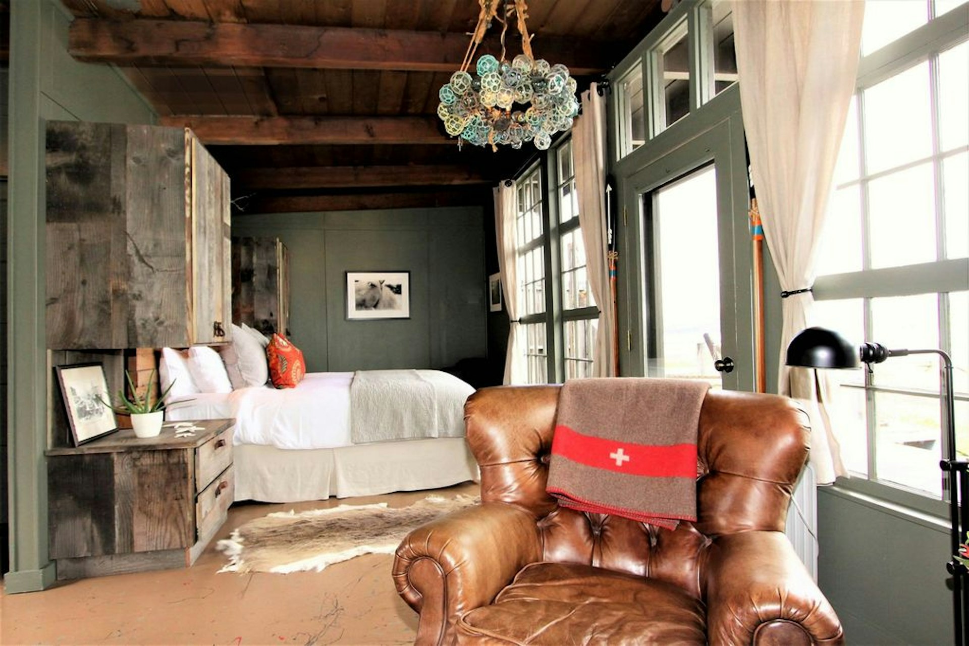 A rustic bedroom with a high beamed ceiling, soft leather armchair in the foreground and a wall of windows on the right that the bed overlooks.