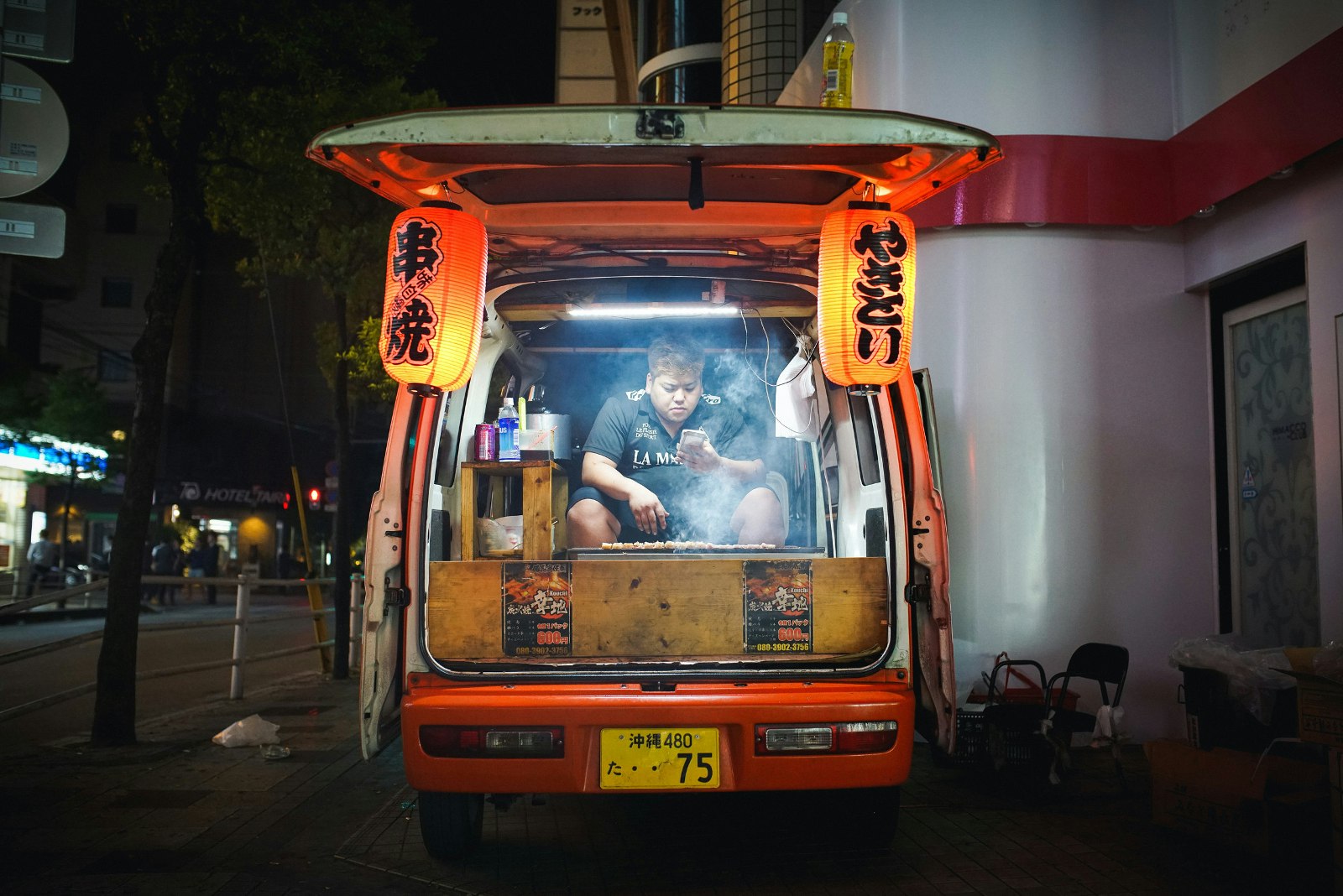 A yakitori vendor is sat in the back of his van behind his griddle while checking his phone in Okinawa, Japan; he is flanked by lanterns on each side hanging from the open hatch door.
