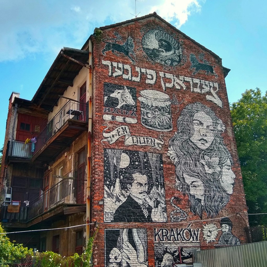 The side of a red brick three-story building shows a black and white mural with Hebrew writing and the prominent image of a woman; Krakow Jewish cultural revival