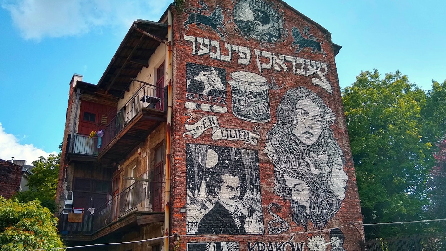 The side of a red brick three-story building shows a black and white mural with Hebrew writing and the prominent image of a woman; Krakow Jewish cultural revival