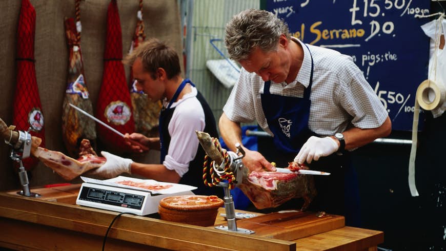 Two market workers in aprons carve jamón directly from the joint at a stall Borough Market