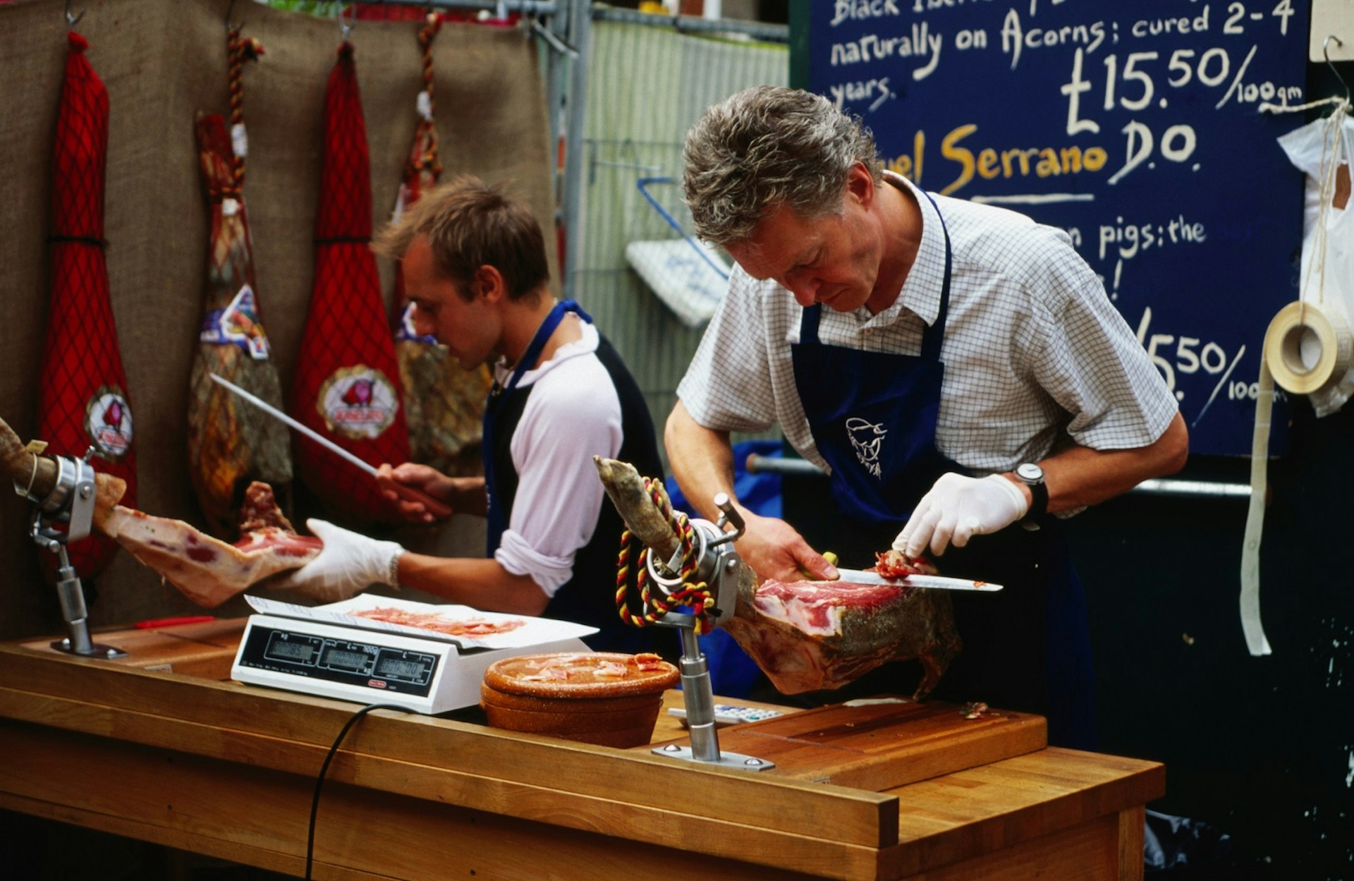 Two market workers in aprons carve jamón directly from the joint at a stall Borough Market