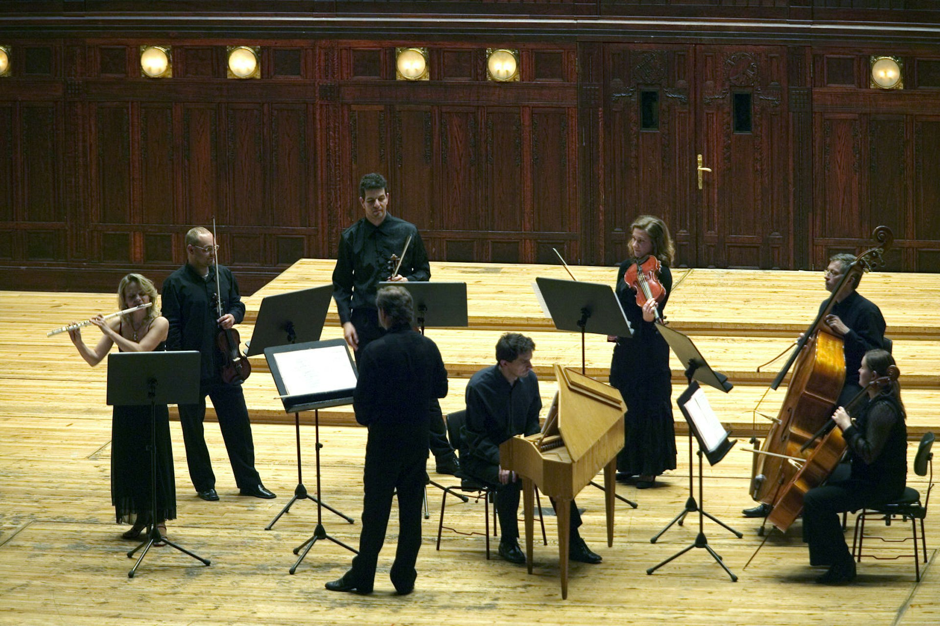 Eight classical musicians, playing flutes, cellos and violins, stand on a wooden stage