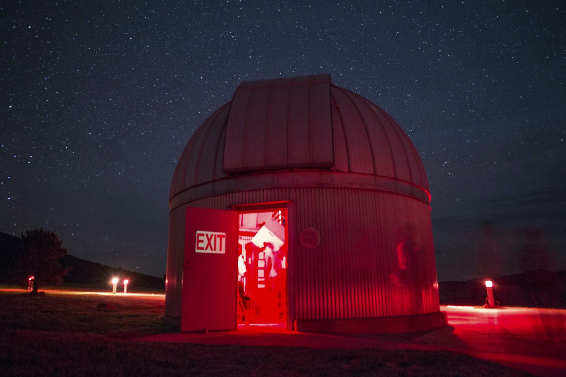 A domed telescope hub at night, lit by red light with the door open, revealing a large 'exit' sign and a blurry interior; Apollo anniversary experiences