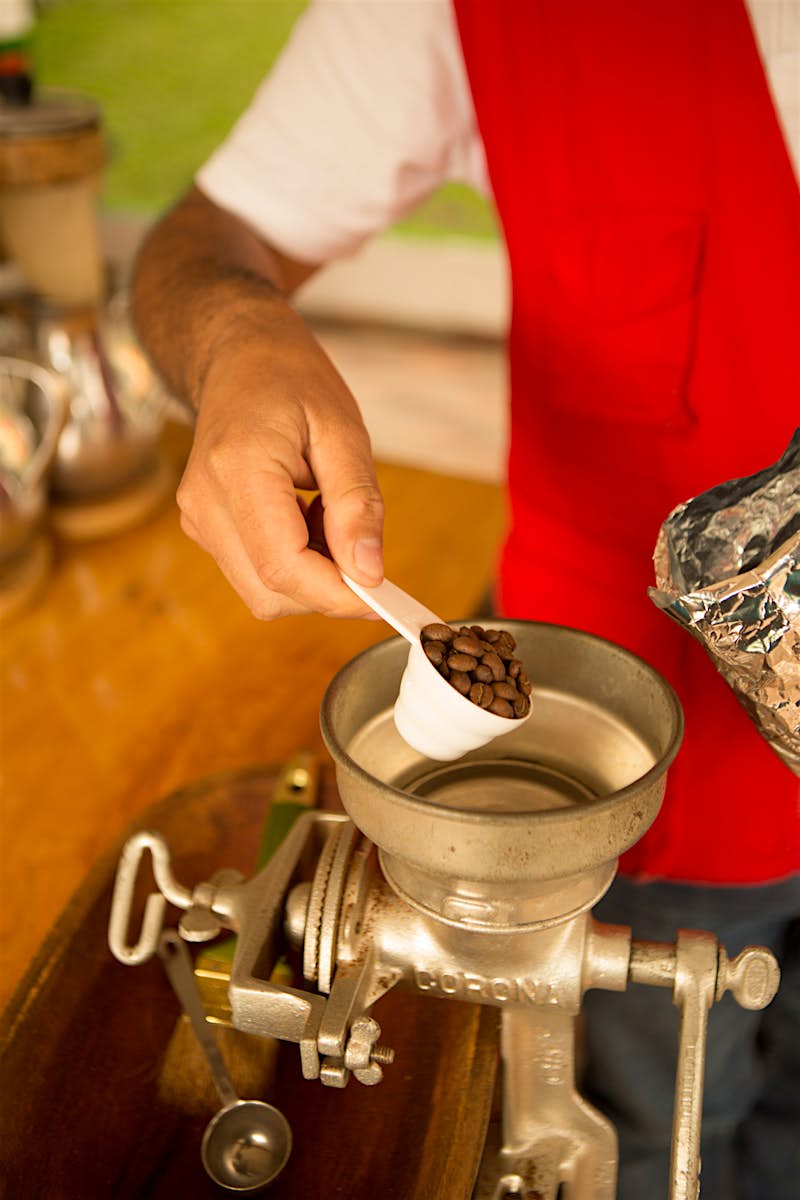 A person pours a tablespoon of coffee beans into a metal coffee grinder to make fresh Colombian coffee. They are wearing a white t-shirt and red vest, the photo is a close up so we can only see their arm and torso.