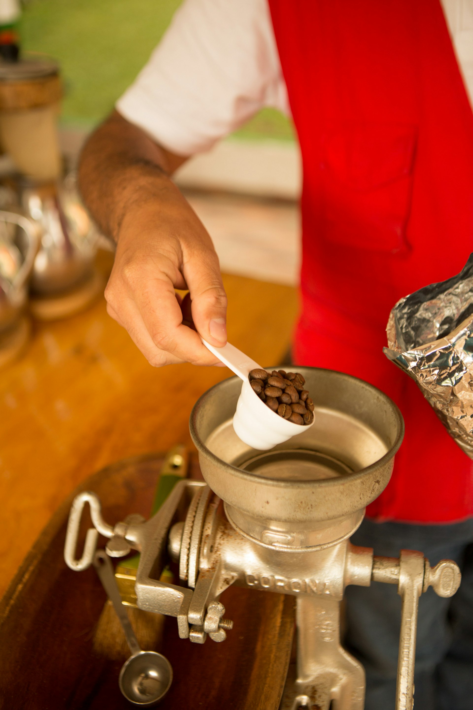 A person pours a tablespoon of coffee beans into a metal coffee grinder to make fresh Colombian coffee. They are wearing a white t-shirt and red vest, the photo is a close up so we can only see their arm and torso.