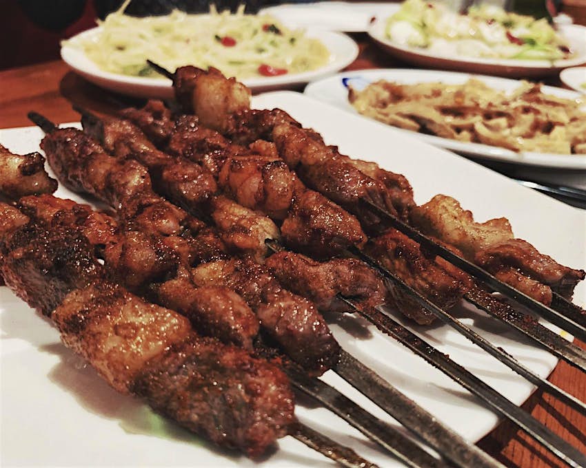 A close up of lamb skewers at Silk Road, Camberwell. Several skewers are piled on a white plate with some other rice dishes in soft focus in the background