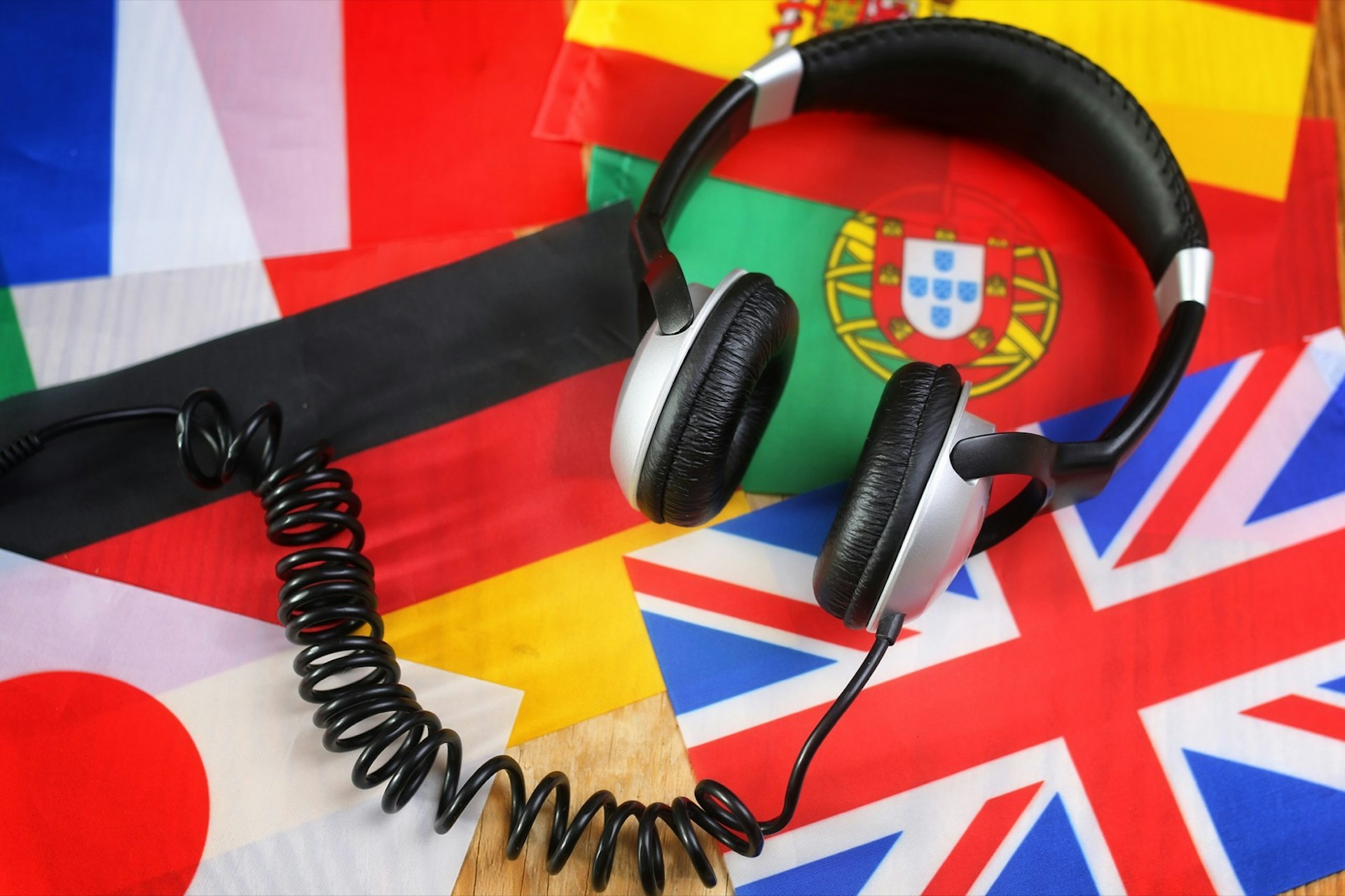 A set of silver and black headphones with a twisty cord sitting on a selection of flags on a wooden table. The flags include Japan, Great Britain, France, Italy, Germany, Portugal and Spain. 