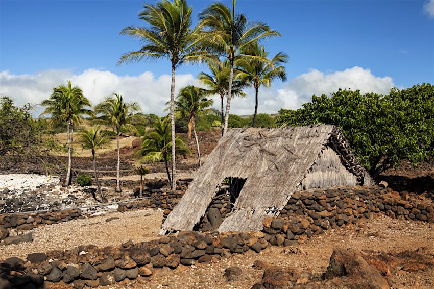 Features - Hawaiian Hale (hut), old Hawaiian Village, Lapakahi State Historical Park, North Kohala Coast, between Kawaihae and Hawi A series of large rocks form a rectangular perimeter around thatch house. There are rock walls and palm trees in the background; free things to do in Hawaii
