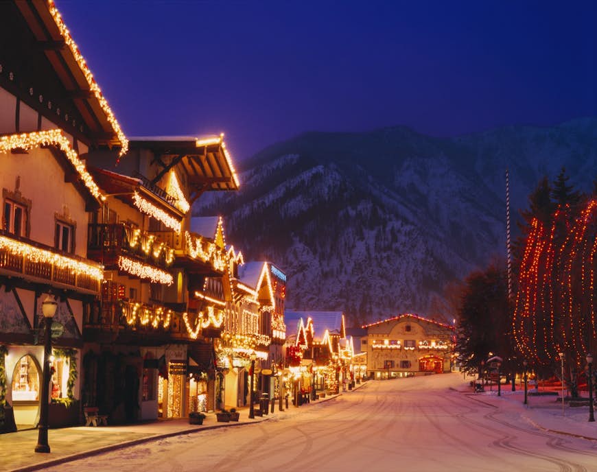 At twilight, glowing strings of lights illuminate a bavarian style village with a snowy street in the foreground. Day trips from Seattle