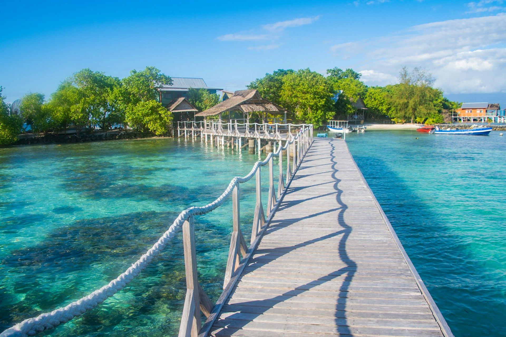 A wooden walkway over crystal-blue waters with a rope railing on one side leads to the Imagination Island resort. The resort is partially obscured by lush greenery, there are a few small boats moored on the shore and an orange building in the far background. 