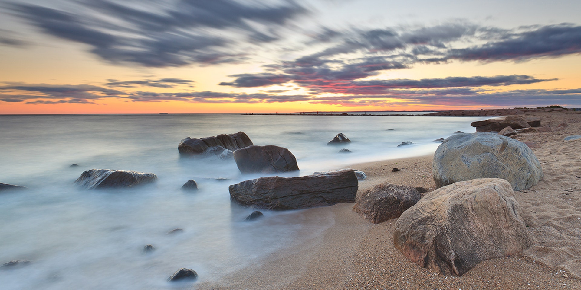 Long exposure of some rocks on a beach, with an orange sunset in the distance; summer weekend trips from New York City