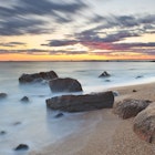 Long exposure of some rocks on a beach, with an orange sunset in the distance;