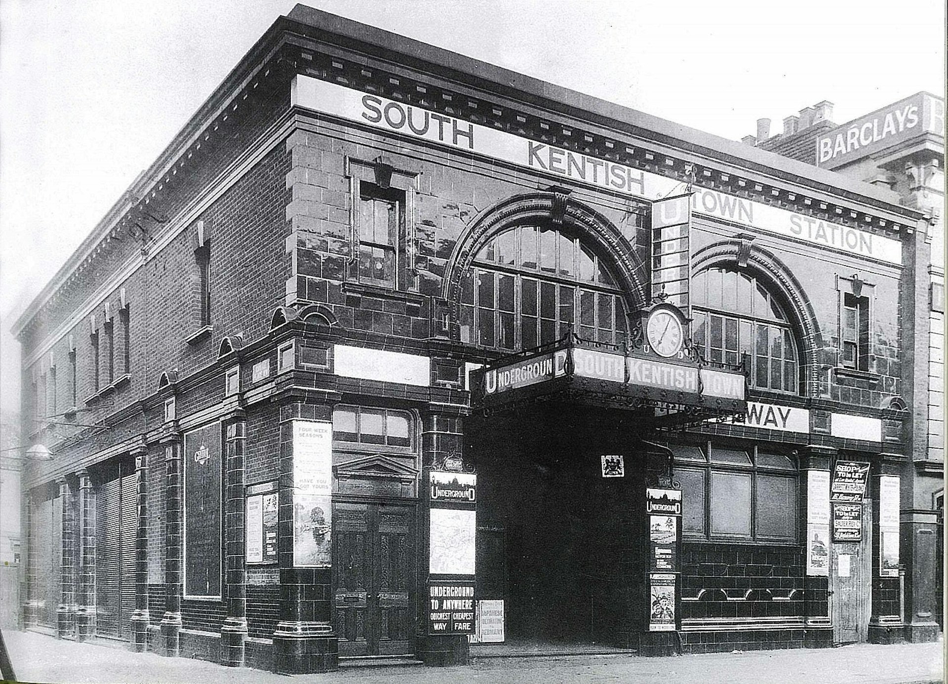 An ageing black and white photo of South Kentish Town tube station exterior; the building is made of bricks with a tiled facade and arched windows over the entrance.