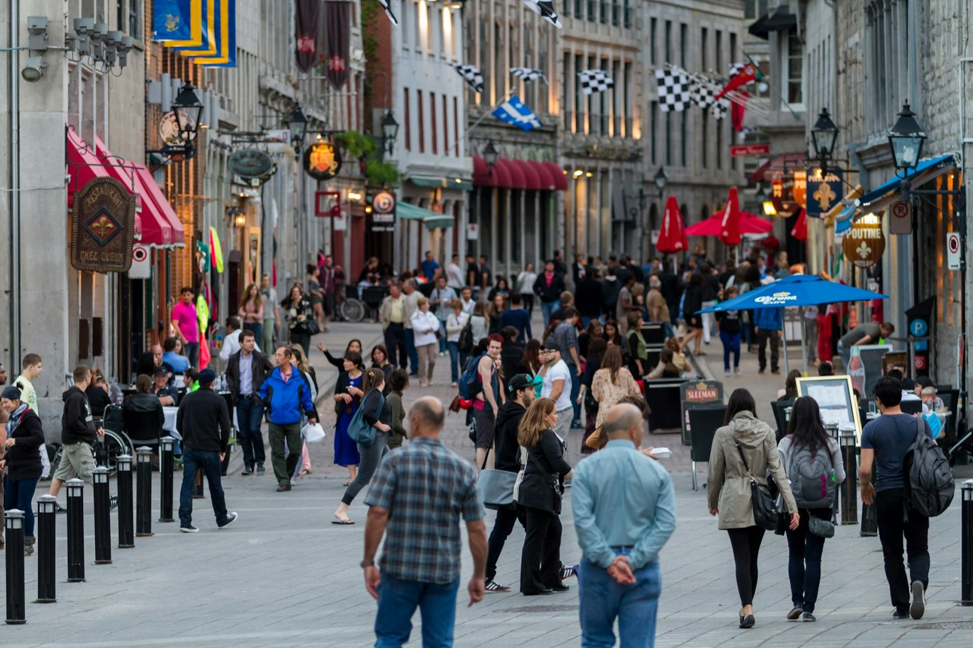 People walk along a cobblestoned street in Montreal with charming shops on both sides and plants, awnings and flags lending color to the gray stone walls; perfect weekend in Montreal