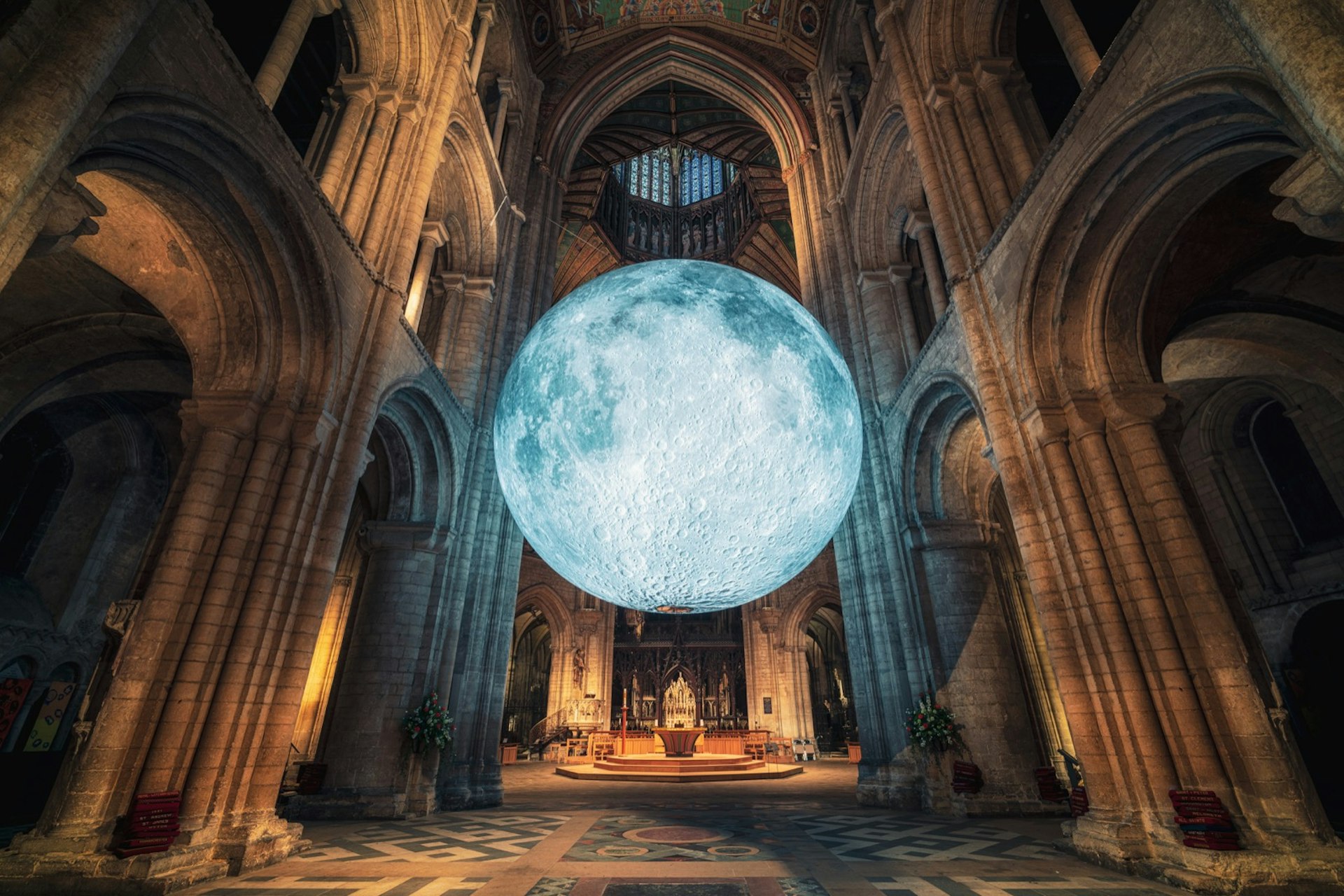 A huge glowing replica of the moon inside the softly lit vaults of a cathedral. The moon appears to be suspended in mid air, surrounded by grey stone columns and stained glass windows. The altar of the church is visible in the background.
