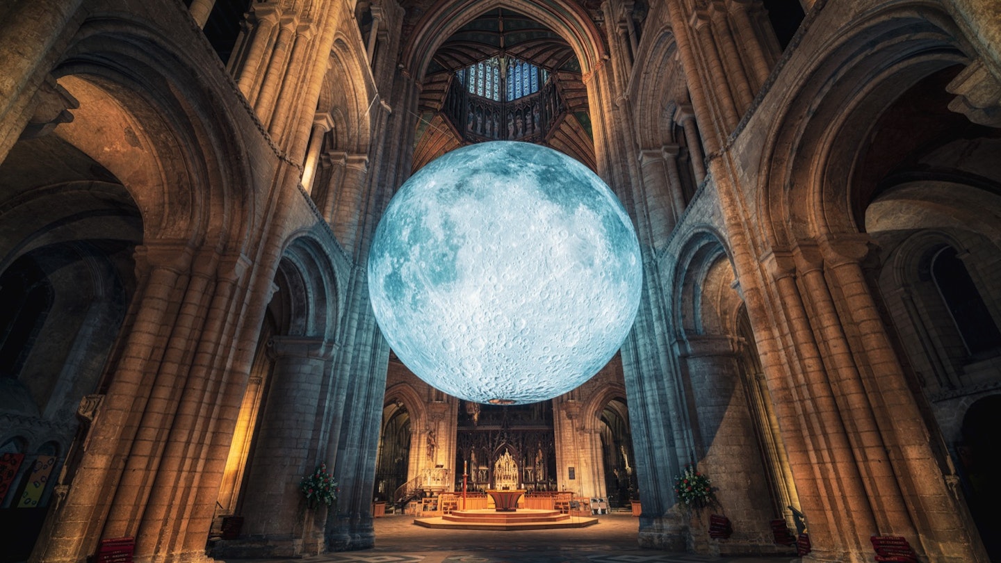 A glowing replica of the moon inside the softly lit vaults of a cathedral